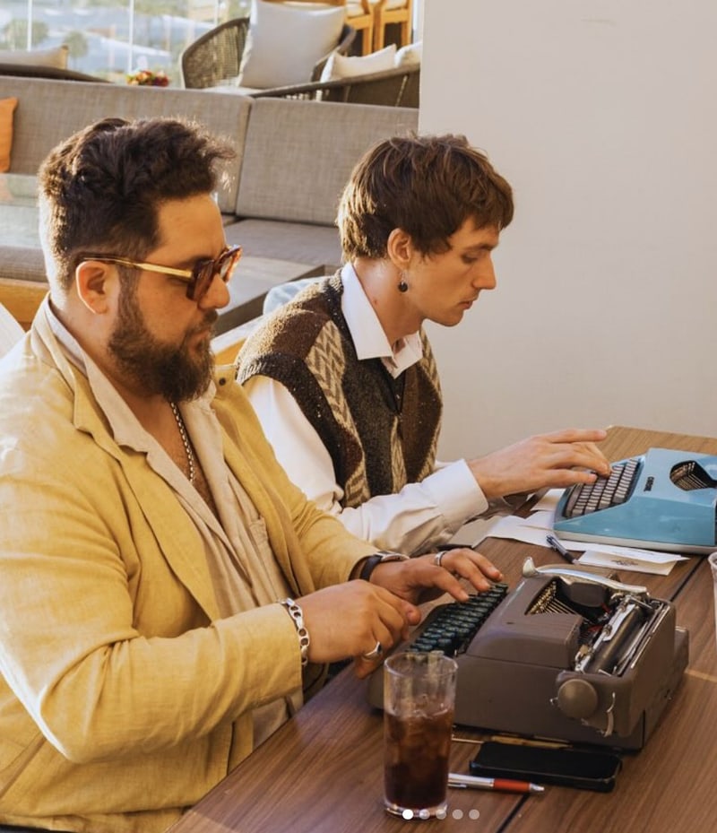 Paasha Motamedi in a yellow linen jacket typing beside a second poet at a blue typewriter