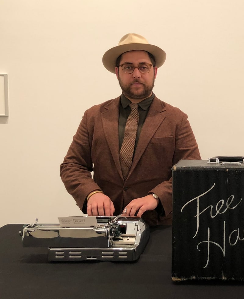 Paasha Motamedi in a cream hat and brown tweed behind a chrome typewriter, a black case lettered Free Haiku beside him