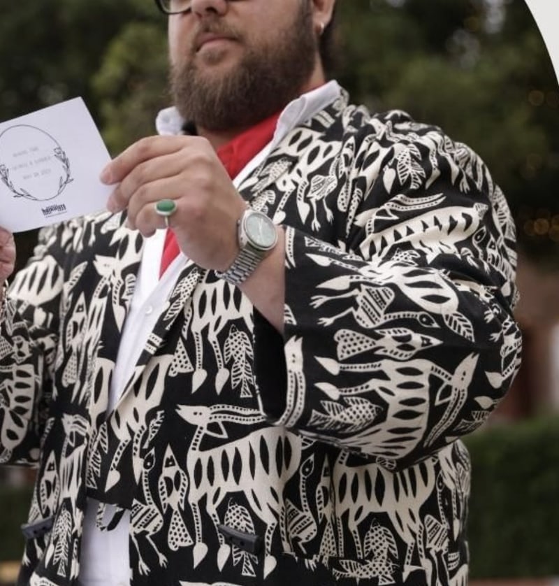 Paasha Motamedi reading a typed poem aloud at an outdoor wedding, in a black and white patterned jacket